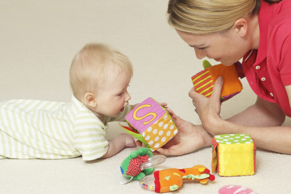 Une dame et un bébé face à face, couchés au sol sur le ventre, jouent à des jeux en tissus.